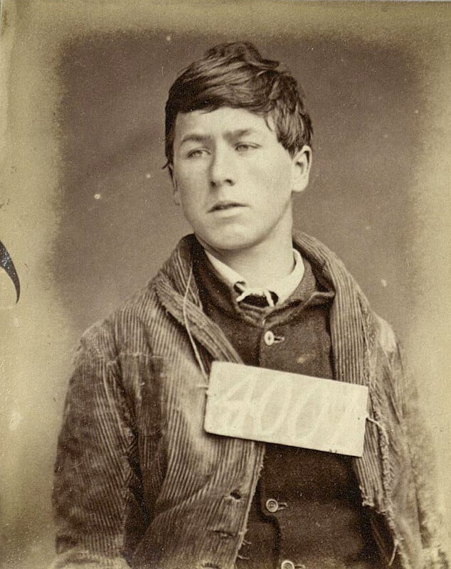 A sepia-toned vintage photograph of a young person wearing a corduroy jacket and a dark shirt, with a rectangular tag displaying a number hanging around their neck. The background is plain, highlighting the individual's expression.