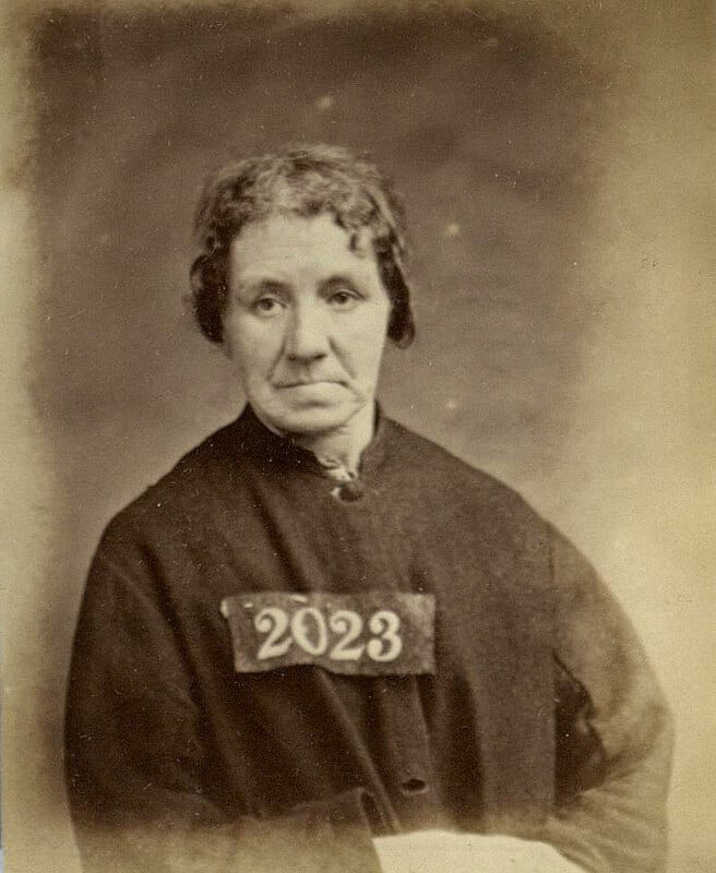 A sepia-toned portrait of an older woman wearing a dark outfit. She has short, wavy hair and a somber expression. A rectangular tag with the number "2023" is pinned to her chest. The background is plain and softly blurred.