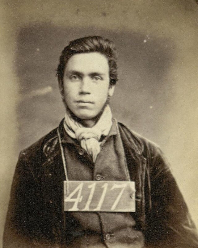 Sepia-toned vintage portrait of a young man wearing a dark jacket, light scarf, and holding a sign with the number 4117. The background is plain and the expression is neutral.