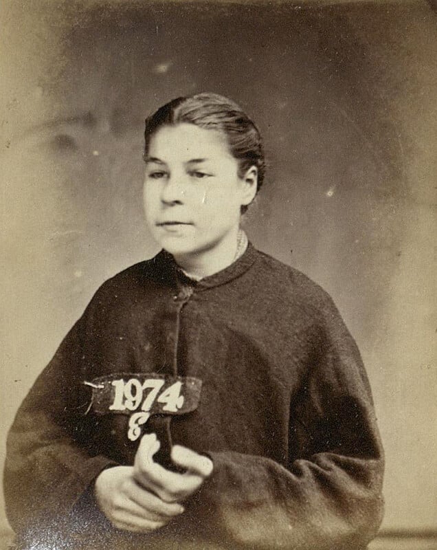 Black and white vintage photograph of a woman with short, dark hair, wearing a dark jacket. She holds a plaque with the number "1974." The background is plain and blurred.