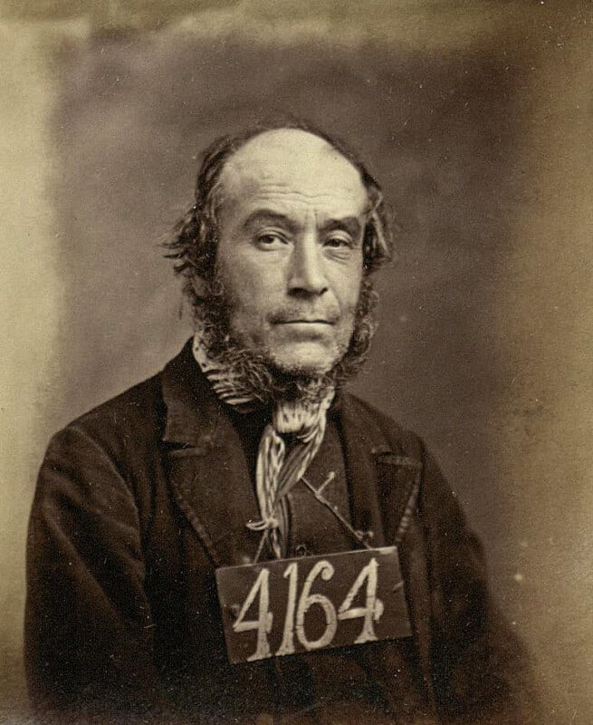 A sepia-toned vintage portrait of a middle-aged man with receding hair and mutton chops, wearing a dark jacket and striped scarf. He has a number "4164" pinned to his chest. The background is plain.