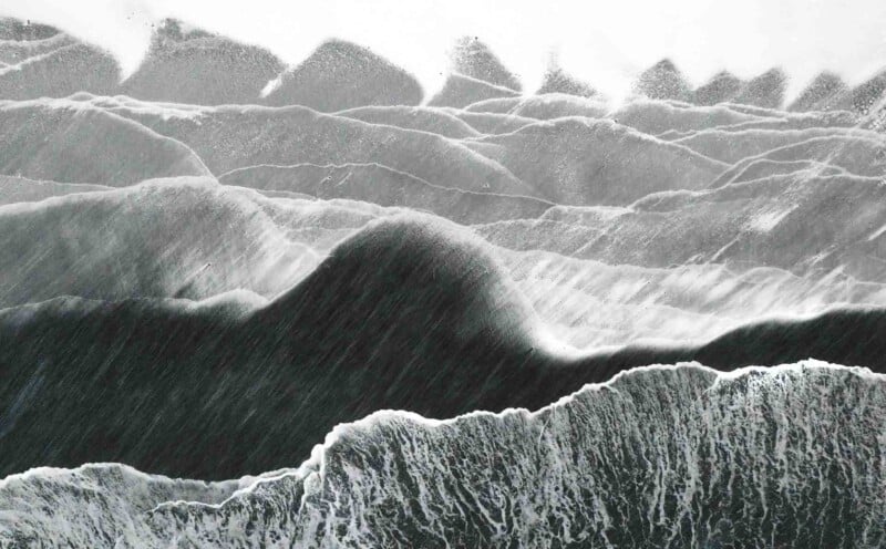 Aerial view of ocean waves crashing with white foam, creating abstract patterns. The water appears dark with contrasting light frothy crests, forming dynamic and textured lines.