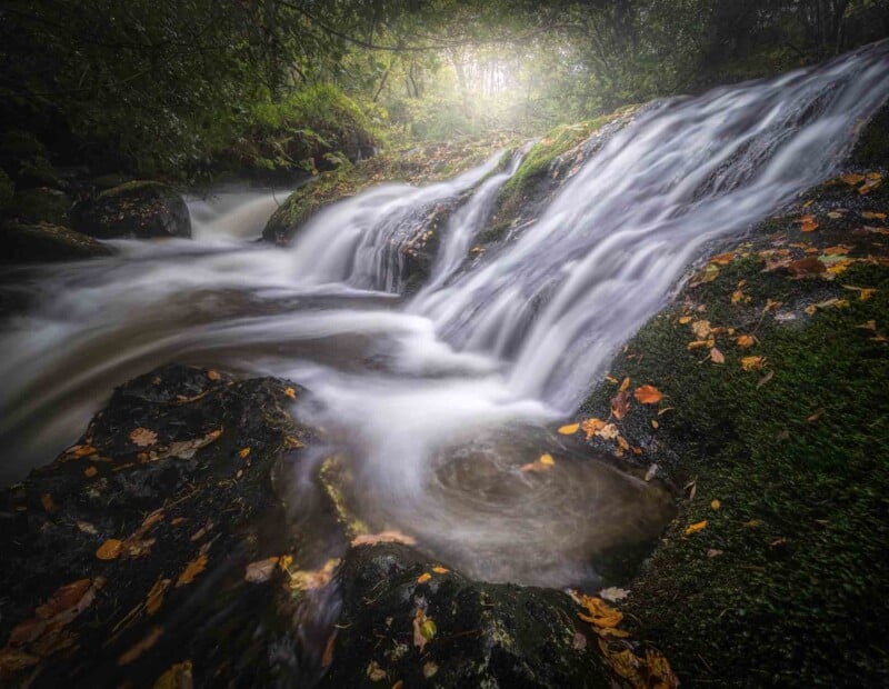 A serene waterfall cascades over moss-covered rocks surrounded by lush green foliage. The water flows smoothly, creating a soft, misty effect, while fallen autumn leaves add a touch of color to the scene. Bright light filters through the trees.