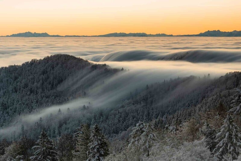 A stunning view of forest-covered hills blanketed in a sea of fog at sunset. The sky features warm orange and yellow hues, creating a serene and mystical landscape with layers of clouds and distant mountain silhouettes.