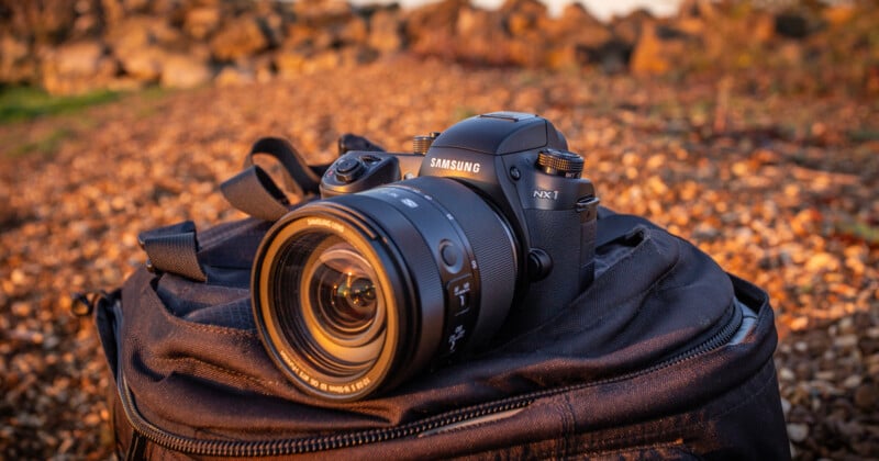 A Samsung NX1 camera with a large lens rests on a black backpack outdoors. The background is a sunlit rocky terrain, creating a warm, natural atmosphere.