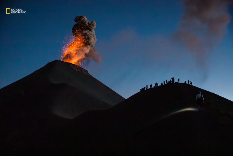 A volcano erupts at dusk, spewing bright orange lava and dark smoke into the sky. Silhouettes of people stand on a nearby ridge, observing the spectacle. The scene is dark, with a faint glow from a flashlight in the foreground.