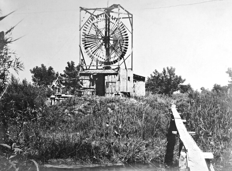 Black and white photo of an early wooden wind turbine on a grassy hill surrounded by vegetation. A wooden walkway leads to the structure, which has a large circular blade within a square frame. Trees are visible in the background under a clear sky.