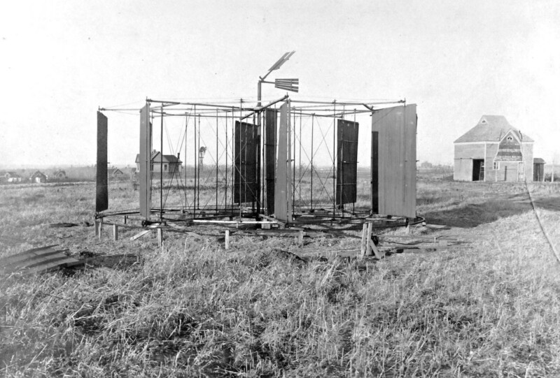 Black and white image of an early experimental wind turbine with large flat panels mounted on a frame in a grassy field. A barn and a few houses are visible in the background.