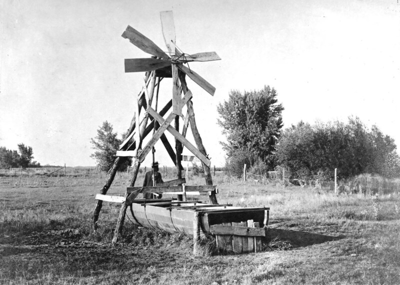 A black-and-white photo of an old windmill with wooden blades standing in an open field. The base is constructed from wooden beams, and there's a trough at the bottom. Trees and a fence are visible in the background.