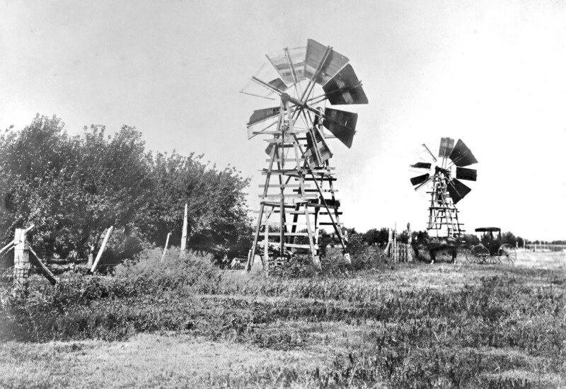 A black and white image of two vintage windmills in a rural landscape. The windmills stand amidst grassy fields and a fenced area with trees in the background. A horse-drawn wagon is visible near the right windmill.