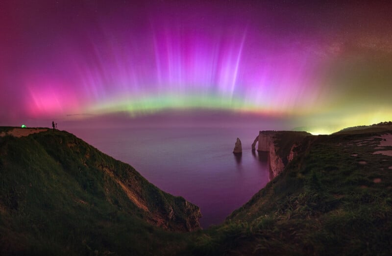 A vibrant aurora displays pink and green lights over a rocky coastal cliff at night. A person stands on the cliff's edge, seemingly photographing the breathtaking scene. The sea reflects the colorful lights under a starry sky.