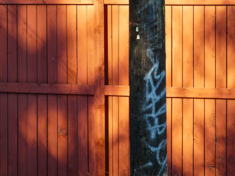 A wooden fence with a warm orange hue is illuminated by sunlight. In front of the fence, a utility pole stands with blue graffiti markings. Shadows of trees are cast on the fence, creating a contrasting pattern.