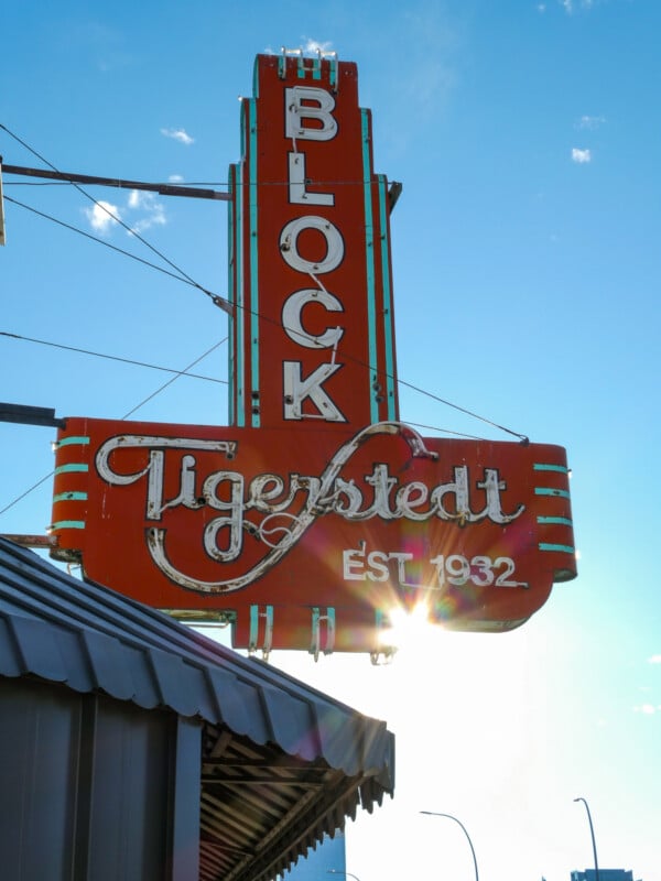 Red vintage sign reading "Block Tigerstedt Est. 1932" with decorative script, mounted on a building facade. The sun shines behind the sign, casting a glow. Blue sky with a few clouds in the background.