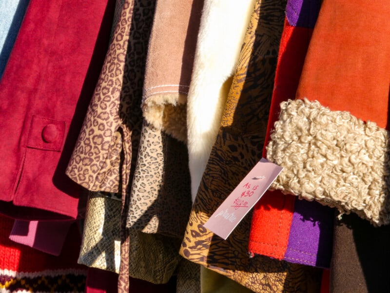 A colorful assortment of clothes is hanging on a rack, featuring various textures and patterns, including leopard print, faux fur, and suede. A pink price tag is visible, displaying "$15 or $30." Bright sunlight highlights the fabric details.