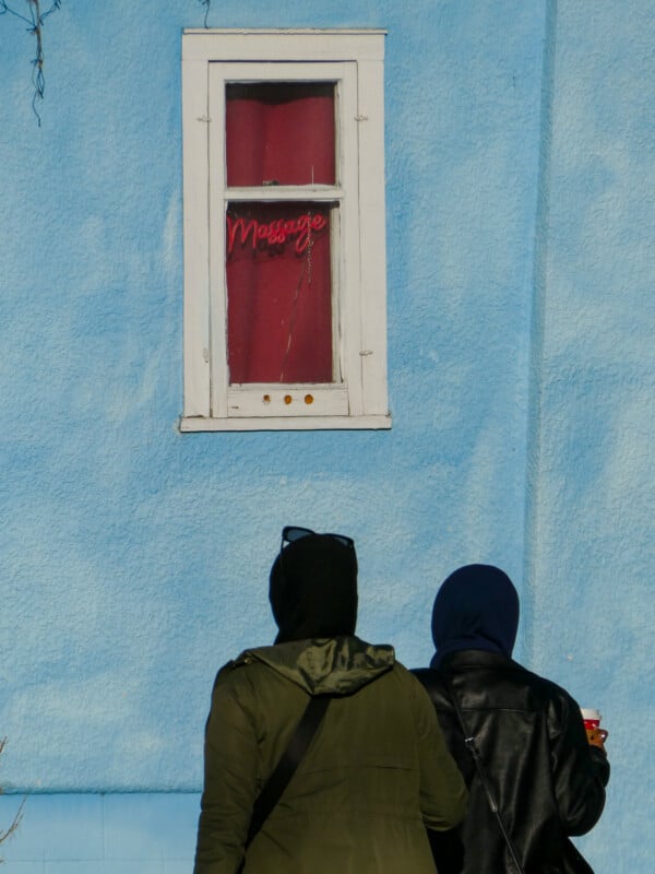 Two individuals stand facing a blue wall with a small window above them. The window is framed in white, with a red curtain and a neon sign that reads "Massage." Both individuals are wearing hooded jackets. One holds a coffee cup.