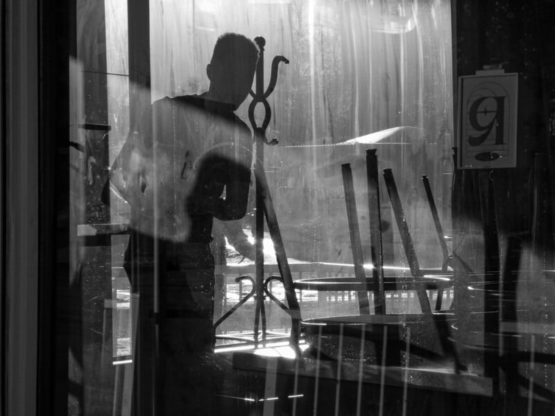 Silhouette of a person seen through a glass door, lifting a chair in a dimly lit room. The reflection creates a layered effect. A sign with the number 9 is visible on the right side. Chairs are stacked on tables in the background.