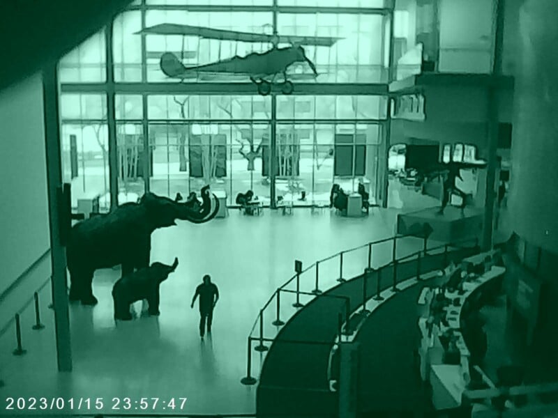 A large indoor museum space with a mammoth and calf statues, an overhead plane model, and people sitting and walking around. The image is tinted in green and shows a reception area on the right. Time stamp reads 2023/01/15 23:57:47.