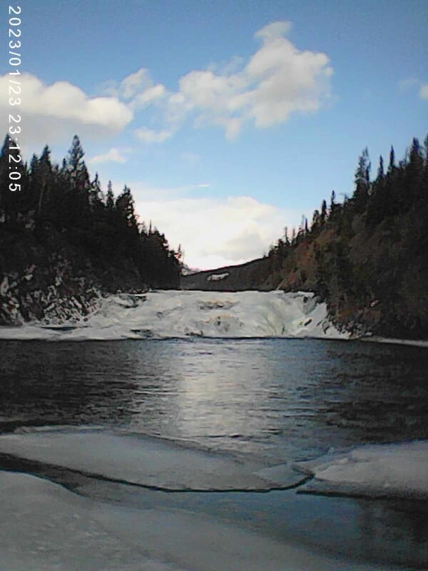 A serene winter landscape features a partially frozen river flowing towards a snow-covered waterfall, surrounded by snow-dusted trees under a blue sky with scattered clouds.