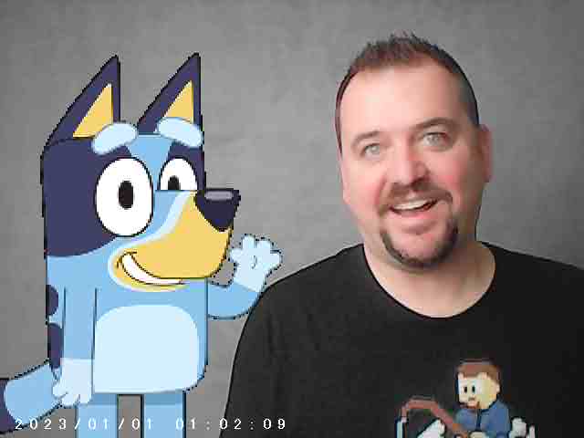 A smiling man stands next to an animated blue dog character with glasses. Both are facing the camera against a neutral backdrop. Timestamp at the bottom: 2023/01/01 01:02:09.