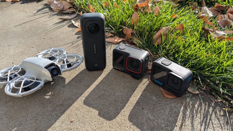 Four action cameras are placed on a concrete surface outdoors with grass and fallen leaves nearby. The devices vary in size and design, including a small white drone camera and black rectangular cameras. The scene is sunny, casting shadows on the ground.