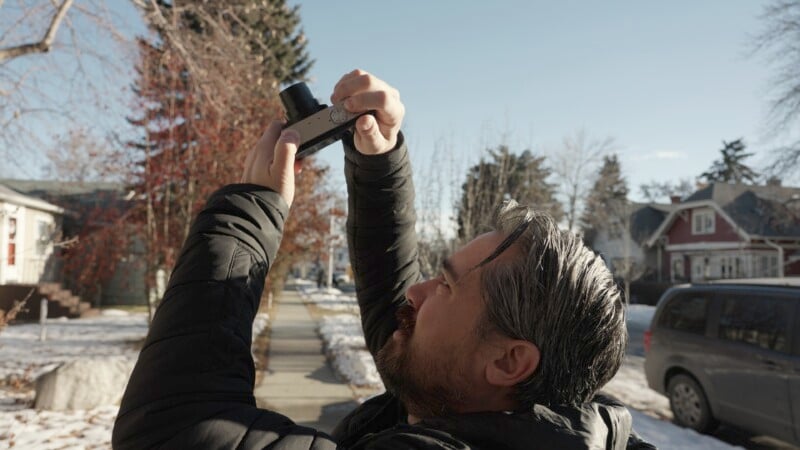 A man in a black jacket holds a camera pointed towards the sky on a sunny winter day. He stands on a snow-dusted sidewalk lined with trees and houses.