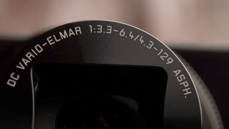 Close-up of a camera lens with the text "DC VARIO-ELMAR 1:3.3-6.4/4.3 12.9 ASPH." engraved on the edge. The lens surface has concentric circular patterns, creating a textured appearance. The background is softly blurred.