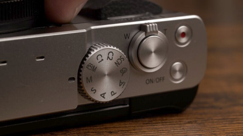 Close-up of a camera's mode dial and power switch. The dial shows options like "M," "A," and "C" for different shooting modes. Next to it is a power switch labeled "ON/OFF" and a zoom button marked "W" and "T". The background is wooden.