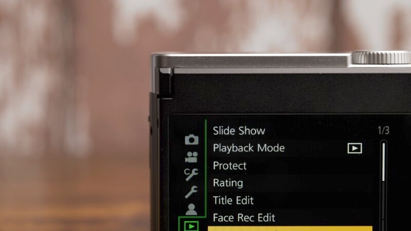 Close-up of a digital camera screen displaying menu options, including Slide Show, Playback Mode, Protect, Rating, Title Edit, and Face Rec Edit. The background is blurred with a warm brown tone.