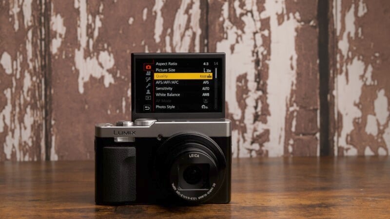 A Panasonic Lumix camera sits on a wooden surface in front of a rustic, weathered wooden wall. The camera's LCD screen is flipped up, displaying its menu interface with various settings options.