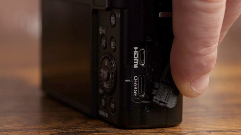 A person plugs a cable into the HDMI and charge port of a black camera. The camera is positioned on a wooden surface, and the person's thumb is visible holding the plug.