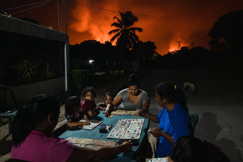 A group of people sit at a table playing a board game outdoors at night. In the background, a large fire lights up the sky, silhouetting palm trees. The scene is dark, with a sense of urgency and tension from the fire.