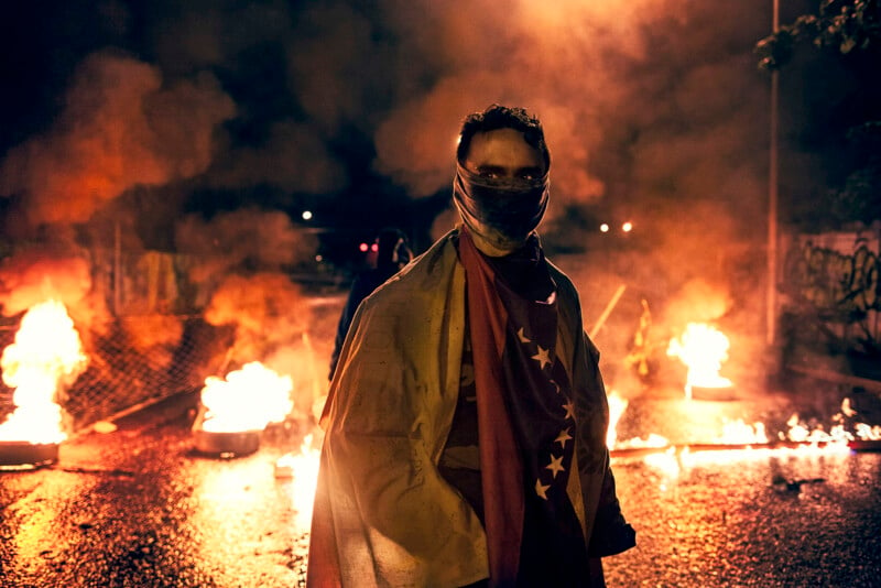 A person stands at night amidst burning barricades on a street. They wear a flag as a cloak and cover their face with a cloth. Smoke fills the air, creating a tense atmosphere.