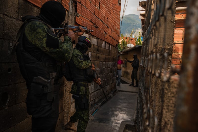 Armed soldiers in camouflage uniforms and masks aim rifles down a narrow alleyway between brick and concrete walls, where a man stands with his hands up. Another soldier is visible further down the alley. A mountain is in the background.