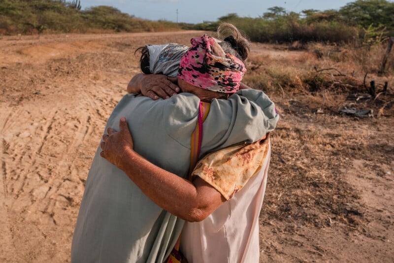 Two people embrace tightly on a dirt road. One wears a headscarf with a pink pattern, and the other has a light-colored garment. The background features dry, sparse vegetation under a clear sky.