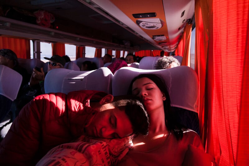 Two people are asleep on a bus, with the person on the left resting their head on the other's shoulder. The bus is filled with passengers, and bright sunlight streams through orange curtains, casting a warm glow on the scene.