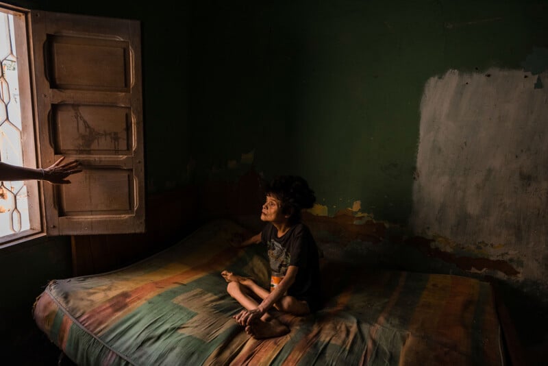 A person sits cross-legged on a worn mattress in a dimly lit room with peeling green walls. Sunlight filters through a partially open window on the left, where a hand is visible, casting light on the person's face.