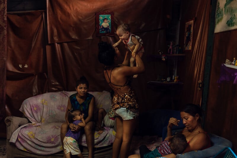 A family sits in a dimly lit room. A woman lifts a baby while another child stands nearby. Two women are seated on a couch; one is breastfeeding another baby. The room is decorated with photos and assorted items.