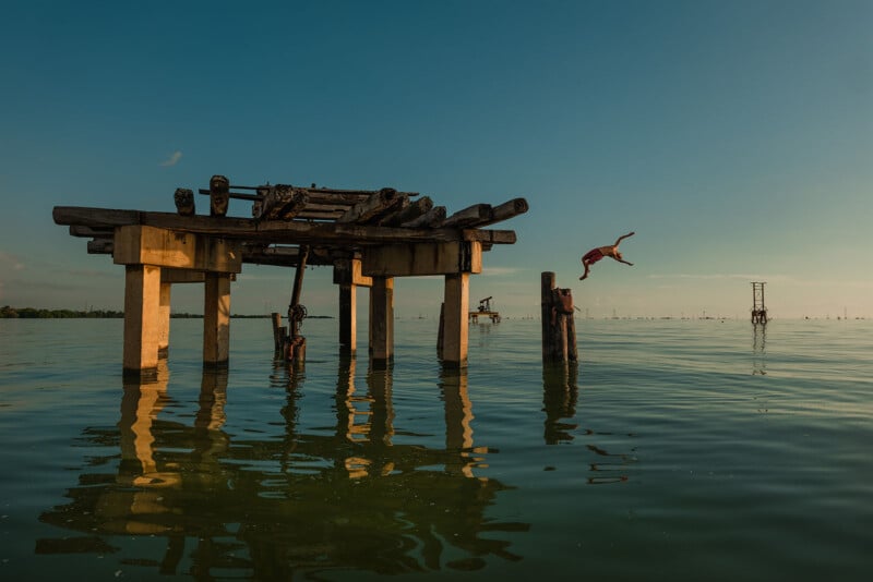 A person is diving off an old wooden structure into calm water during sunset. The sky is clear, and the structure casts a reflection on the water's surface.