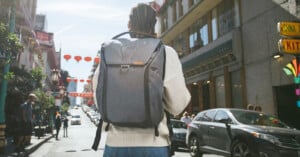 Person with a gray backpack walking down a busy city street lined with vehicles and pedestrians. The street has red lanterns hanging above, and signs in Chinese characters are visible on buildings. The person wears a white sweater and jeans.