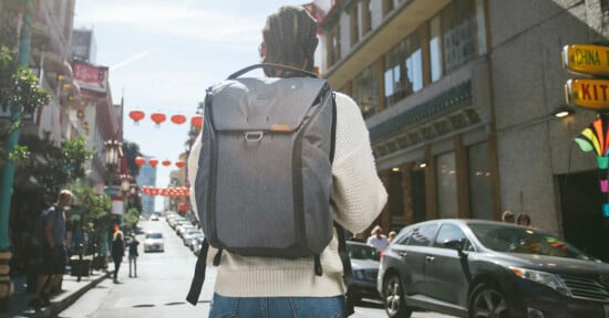 Person with a gray backpack walking down a busy city street lined with vehicles and pedestrians. The street has red lanterns hanging above, and signs in Chinese characters are visible on buildings. The person wears a white sweater and jeans.