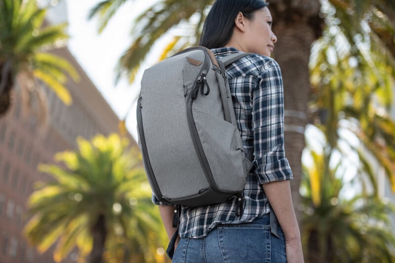 A person wearing a gray backpack and a plaid shirt stands outdoors, with tall palm trees and a building in the background. The sky is clear and sunny, suggesting a warm climate.