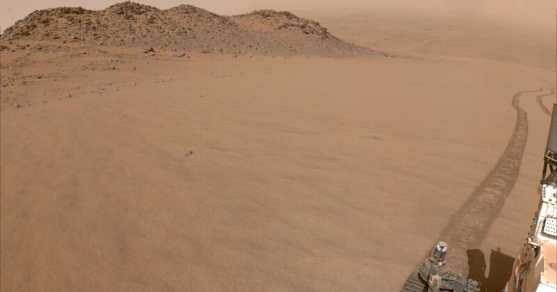 A dusty Martian landscape with rolling hills in the background. Tire tracks from a rover are visible on the right side, leading from the foreground into the distance. The sky appears hazy and the terrain is covered in fine reddish sand.