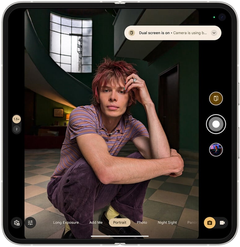 A person with red hair crouches indoors, facing the camera, holding their head with one hand. They wear a striped shirt and purple pants. A window and curved stairway are in the background. The image is displayed on a foldable device screen.