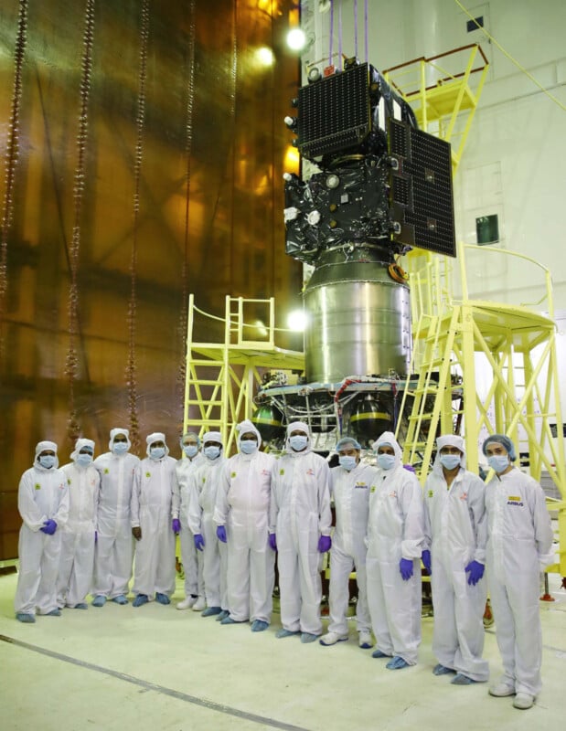 A group of people in white protective suits and masks stand in front of a large, complex piece of machinery in an industrial setting. The machinery is surrounded by a yellow platform with stairs.