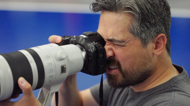 A bearded man with gray hair is intently looking through the viewfinder of a large camera with a long lens, appearing focused. The background is blurred in blue tones.