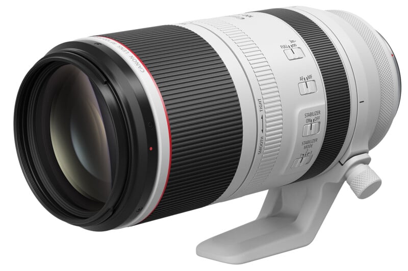 A Canon telephoto camera lens with a white and black barrel, featuring a red ring near the front. It has multiple switches for settings and comes with a tripod mount collar.