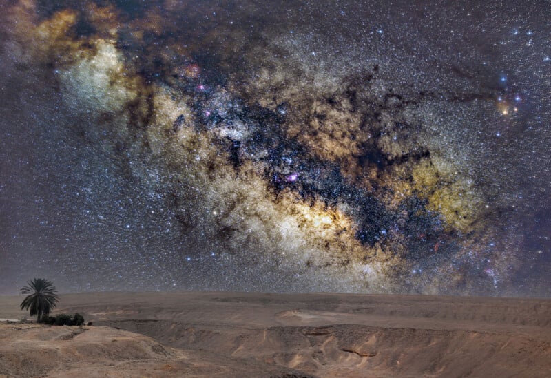 A vast desert landscape with a lone palm tree at the horizon under a vibrant, star-filled night sky. The Milky Way galaxy is prominently visible, with colorful nebulae and dense clusters of stars creating a striking cosmic display.