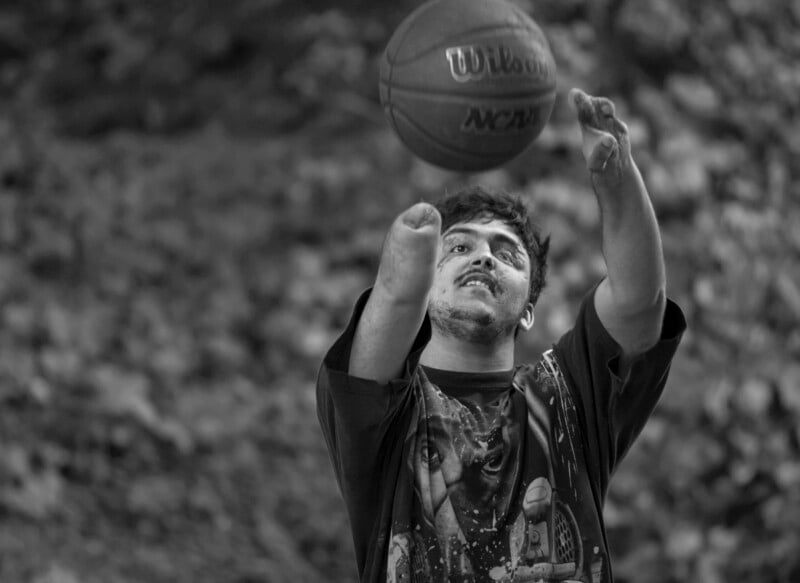 A person with a limb difference plays basketball outdoors, focusing on the ball as they throw it upwards. The image is in black and white, capturing movement and determination.