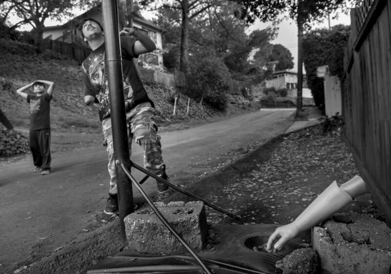 Two boys stand on a quiet street, one examining a pole, while the other watches. Nearby on the ground, a doll's arm protrudes from a concrete block. The scene is surrounded by trees and houses.