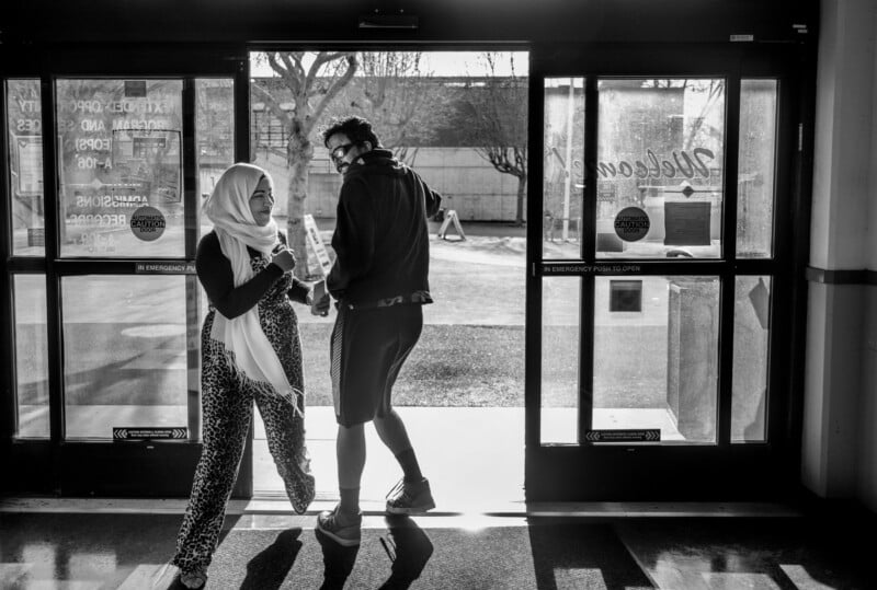 Two people exit a building through automatic doors. The woman on the left, wearing a headscarf and patterned pants, smiles while holding a drink. The man on the right, in shorts, carries a bag and looks back over his shoulder. Sunlight streams in.